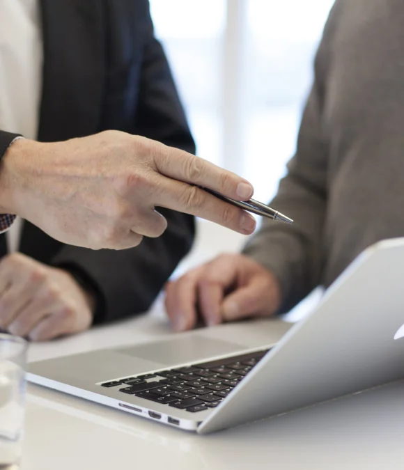 Two IT professionals discussing information displayed on a laptop screen, with one person pointing at the screen using a pen during a collaborative meeting.