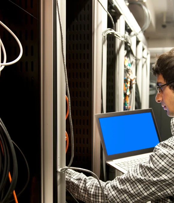 IT technician working in a server room, connecting network cables while using a laptop with a blue screen for diagnostics.
